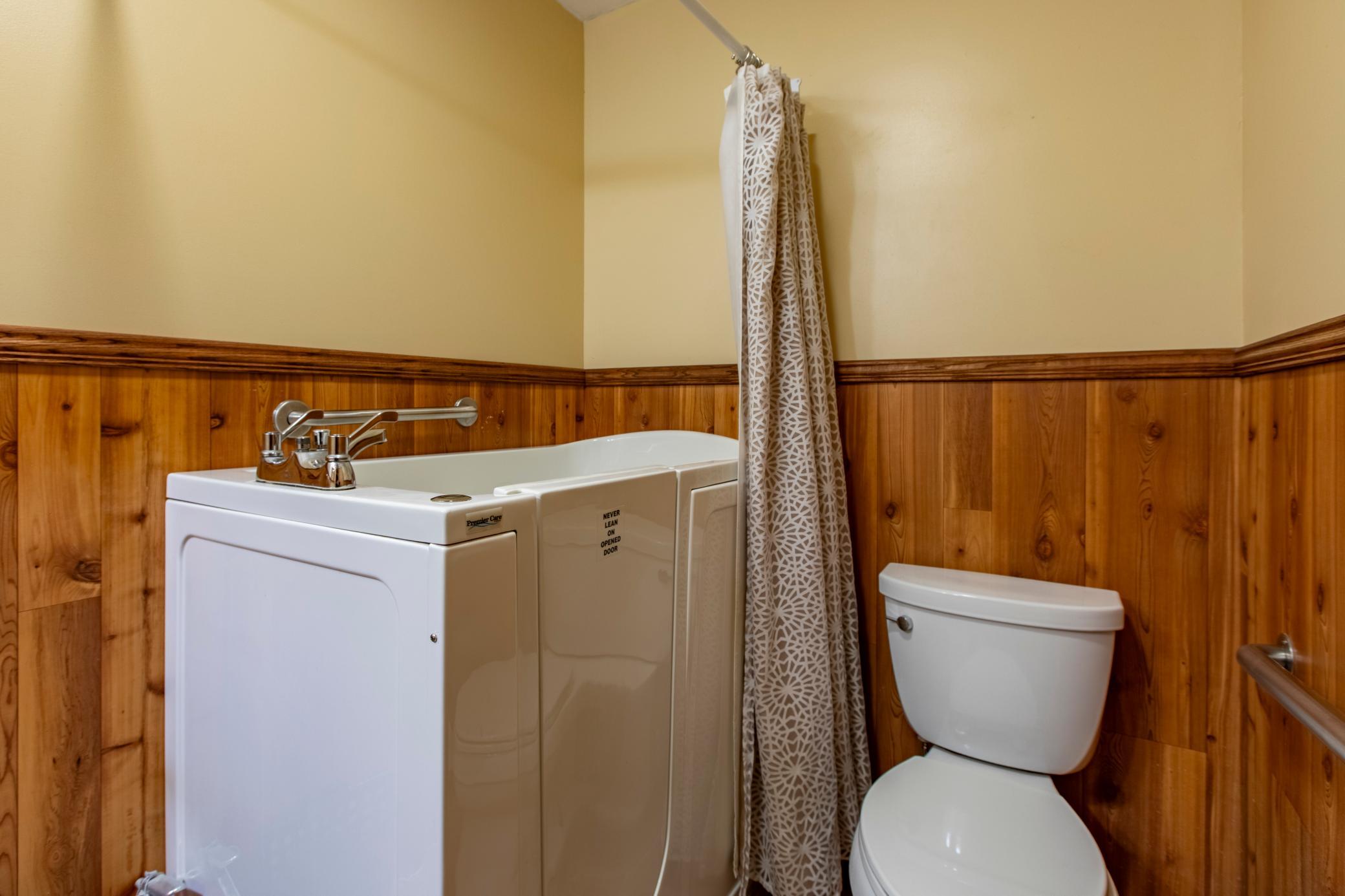 Bathroom with a white walk-in tub, toilet, patterned shower curtain, and wood-paneled walls.