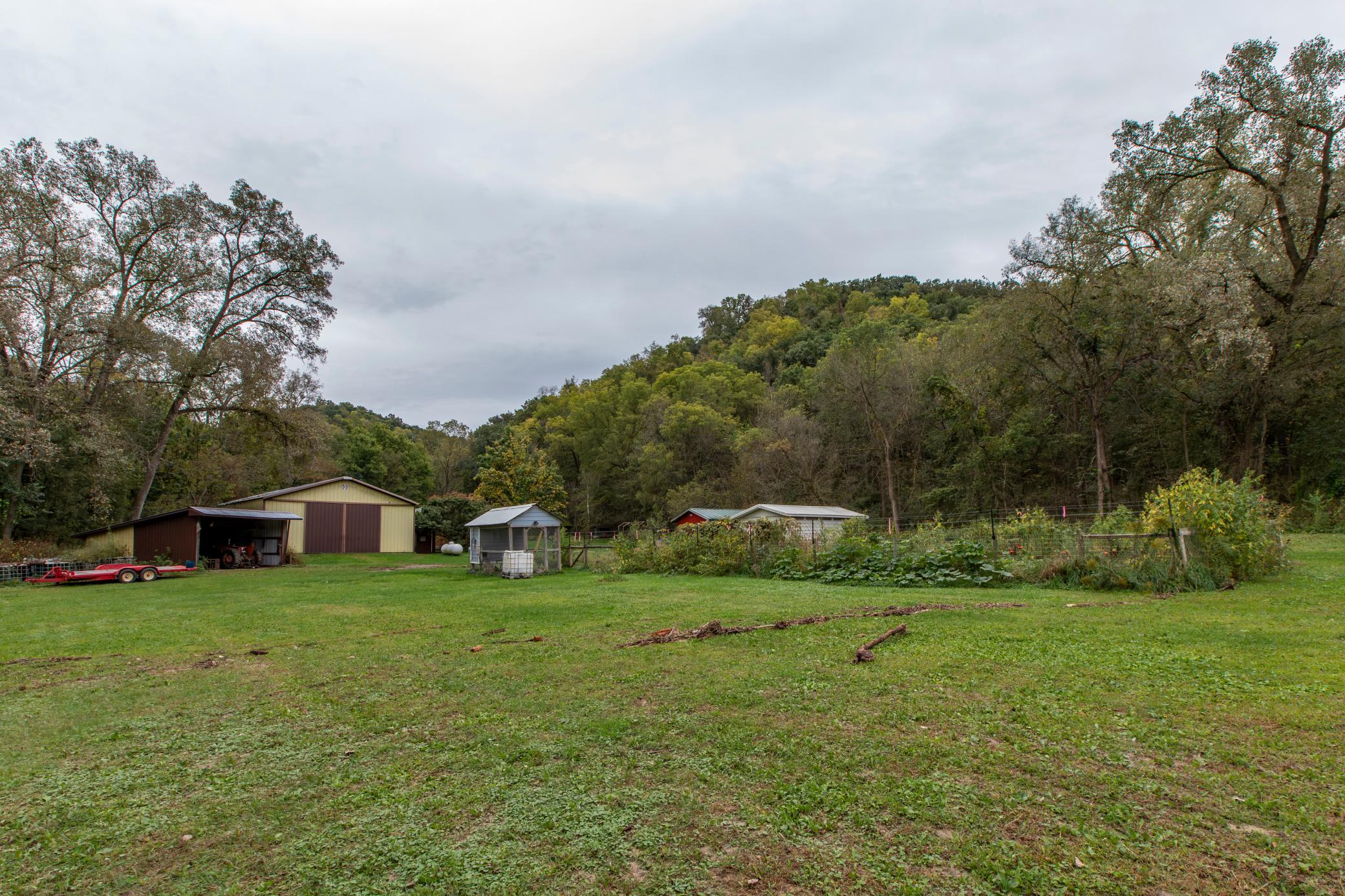Rural property yard with several outbuildings, a fenced garden, and wooded hills.