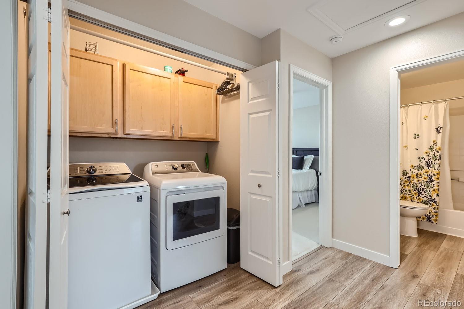 Laundry room with washer, dryer, and light wood cabinets; bedroom and bathroom are visible through open doorways.