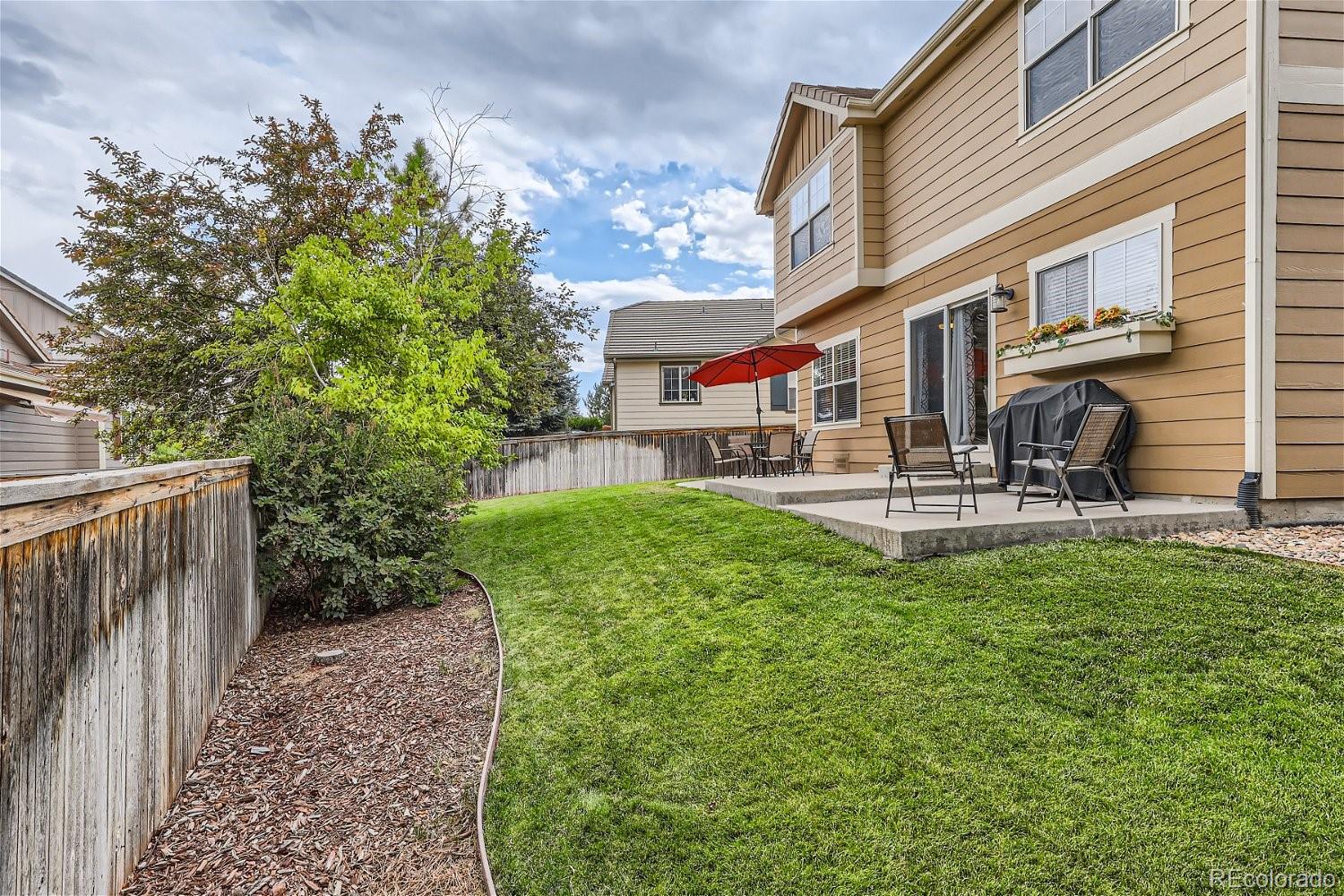 Backyard lawn and patio with outdoor seating, a grill, and red umbrella beside a tan house.