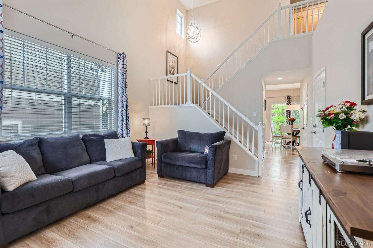 Furnished living room with vaulted ceiling, dark seating, light wood floors, and an open staircase.