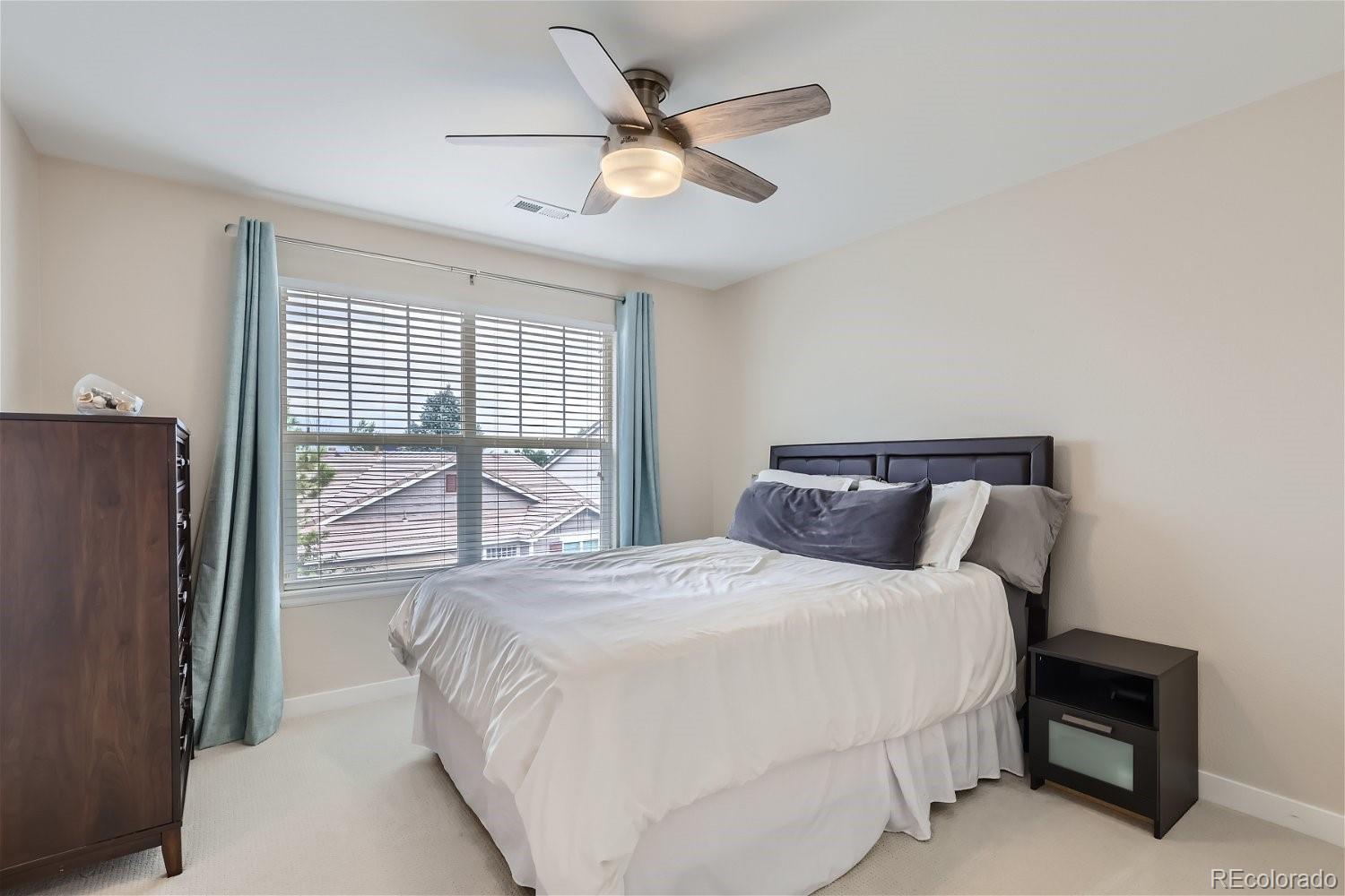 Furnished bedroom with a white bed, ceiling fan, and window framed by blue curtains.