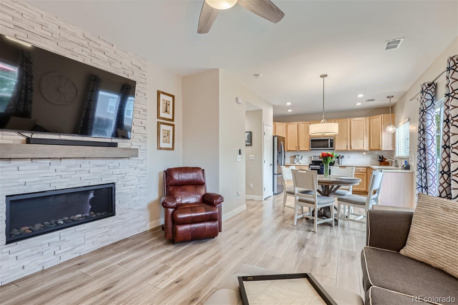 Open-plan living room and kitchen with a stacked-stone fireplace wall, leather recliner, and light wood cabinets.