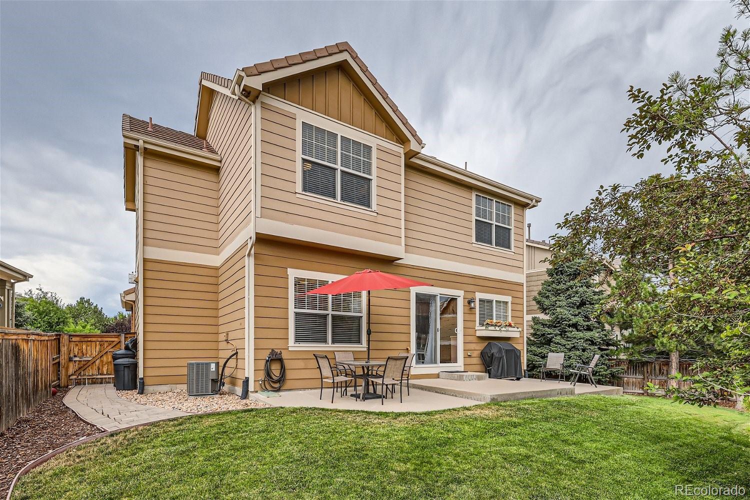 Two-story tan home with a red patio umbrella, outdoor dining set, and fenced grassy backyard.