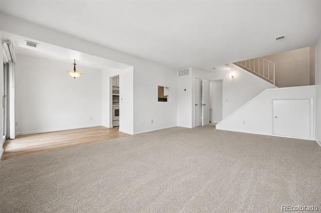 Vacant living room with beige carpet, staircase, and a glimpse of the kitchen.