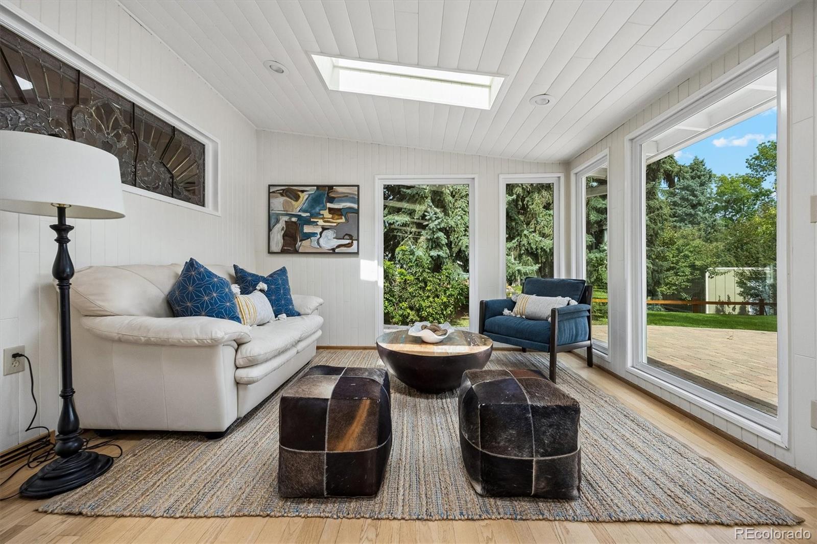 Sunroom with a skylight, white sofa, blue armchair, and large windows overlooking trees and a patio.