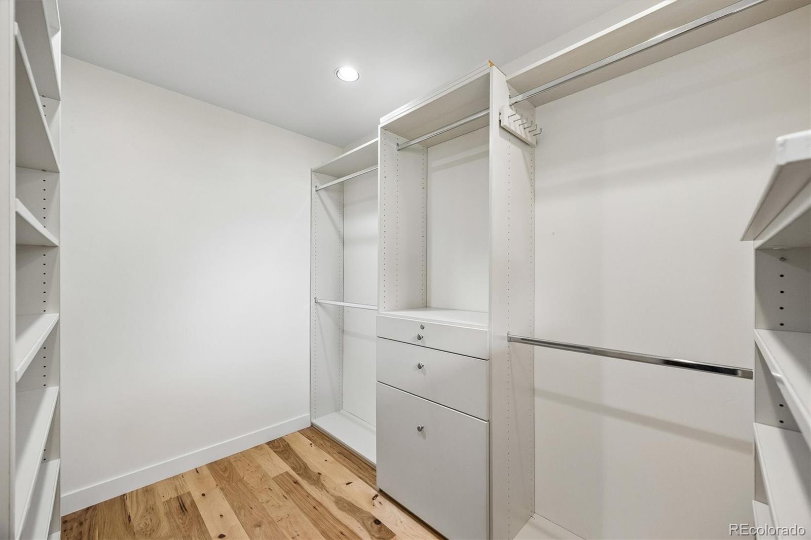 Walk-in closet with white built-ins, hanging rods, central drawers, and light wood flooring.