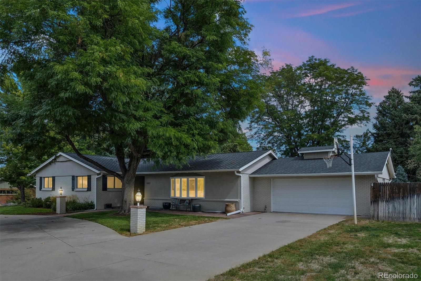 Single-story gray brick home with a two-car garage, mature trees, and a basketball hoop at dusk.