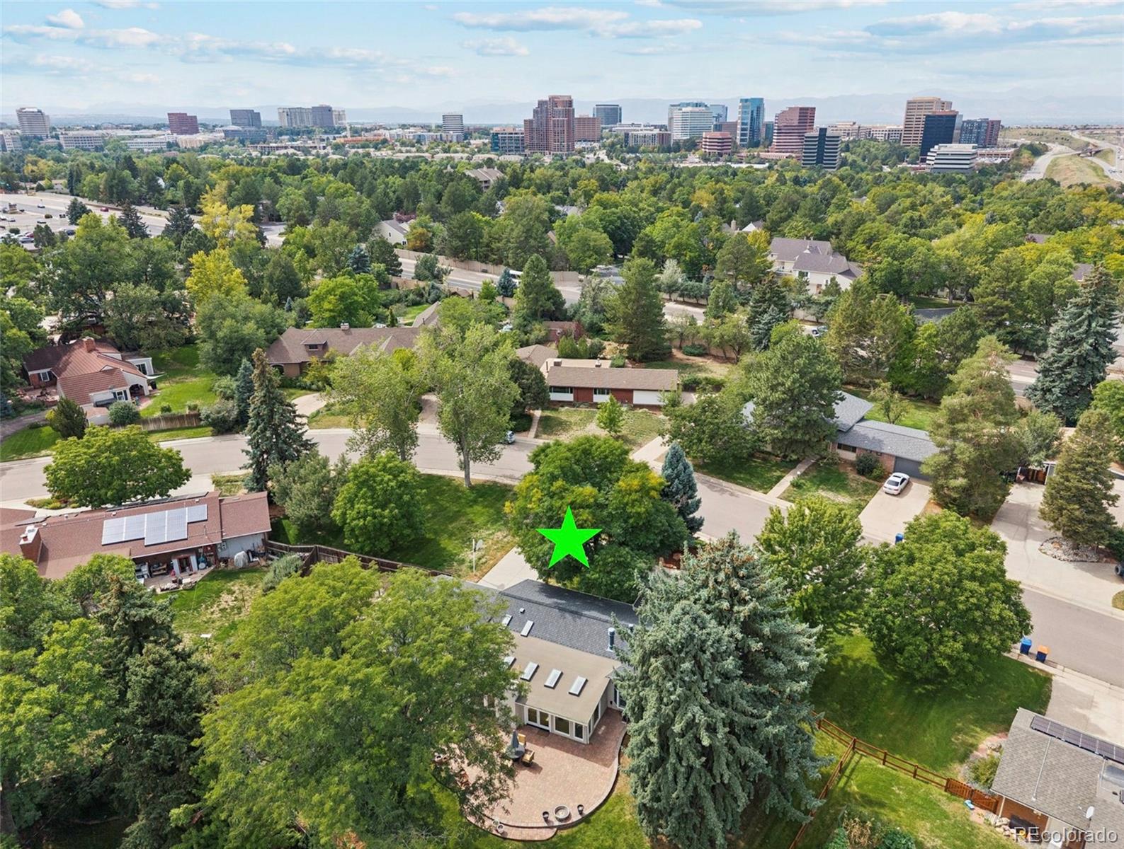 Aerial view of a tree-covered residential lot marked by a green star, with homes and a distant city skyline.