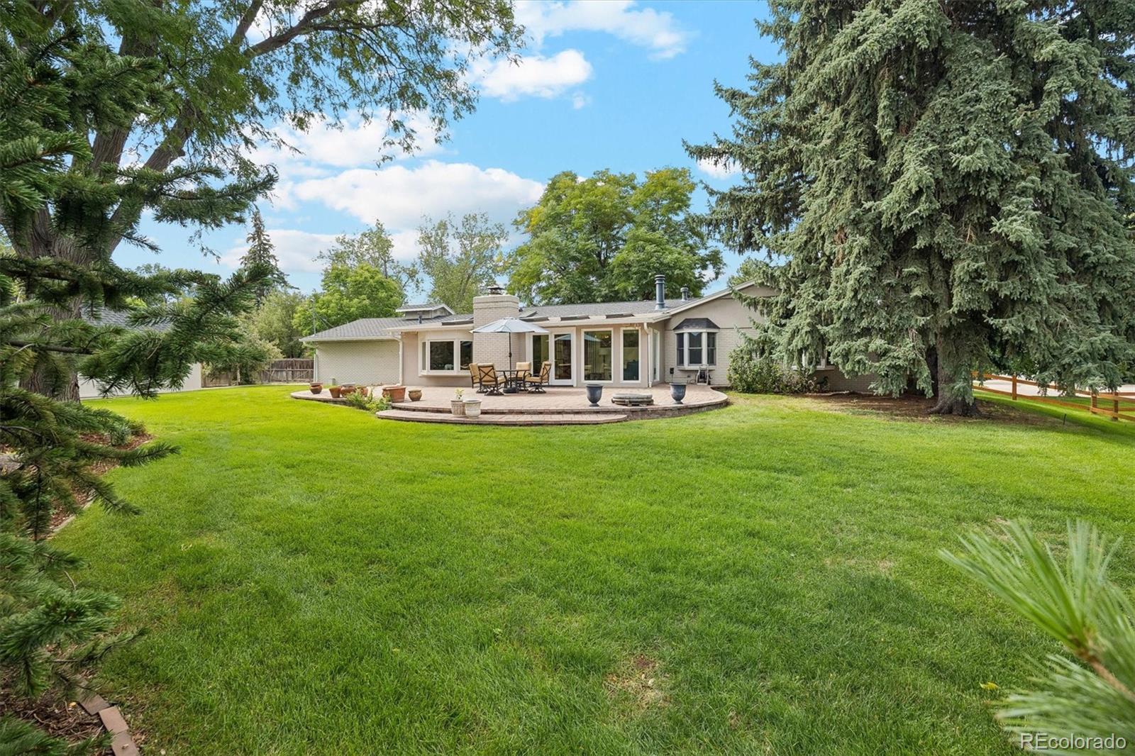 Rear of a single-story home with a curved patio, outdoor dining set, and expansive lawn beneath mature trees.