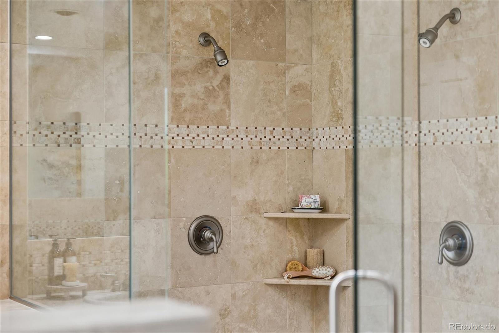 Walk-in shower with beige stone tile, mosaic accent band, glass enclosure, and built-in corner shelves.