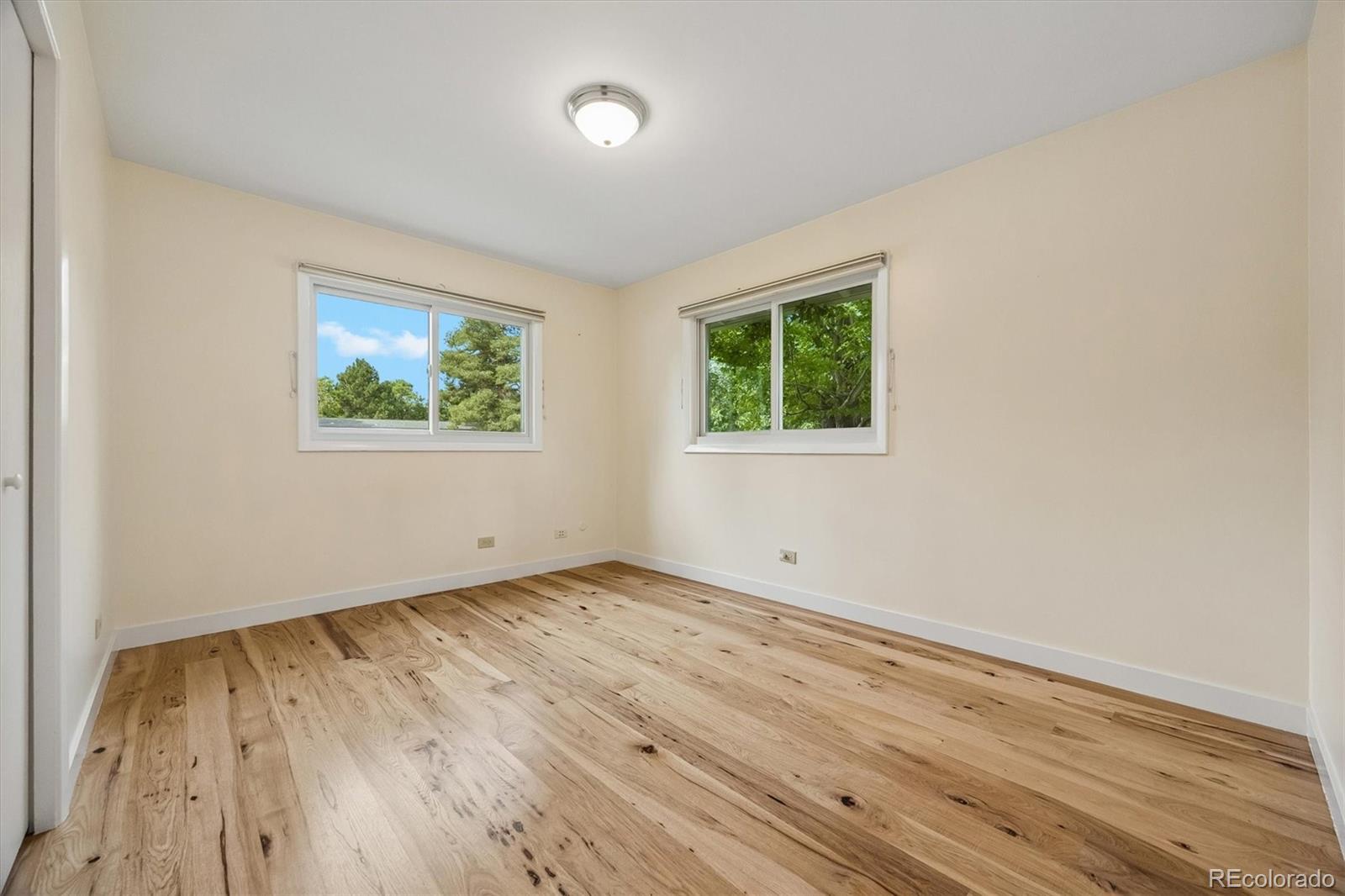 Empty bedroom with natural-wood plank floors and two windows overlooking trees.