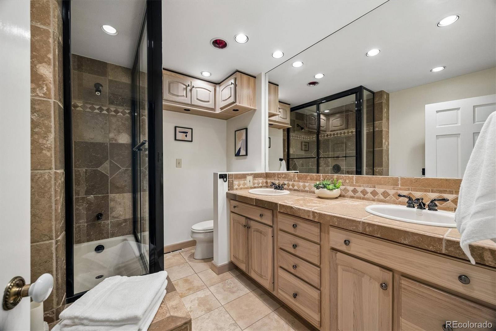 Bathroom with a double-sink vanity, tan stone surfaces, and a black-framed glass shower.