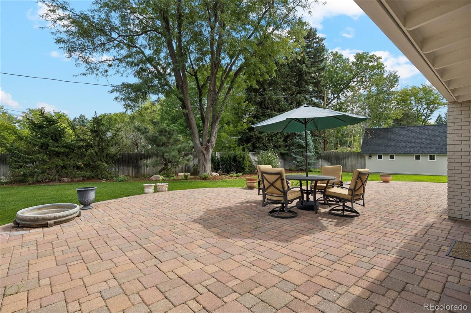 Brick-paver backyard patio with outdoor dining set and umbrella beside a lawn and mature trees.
