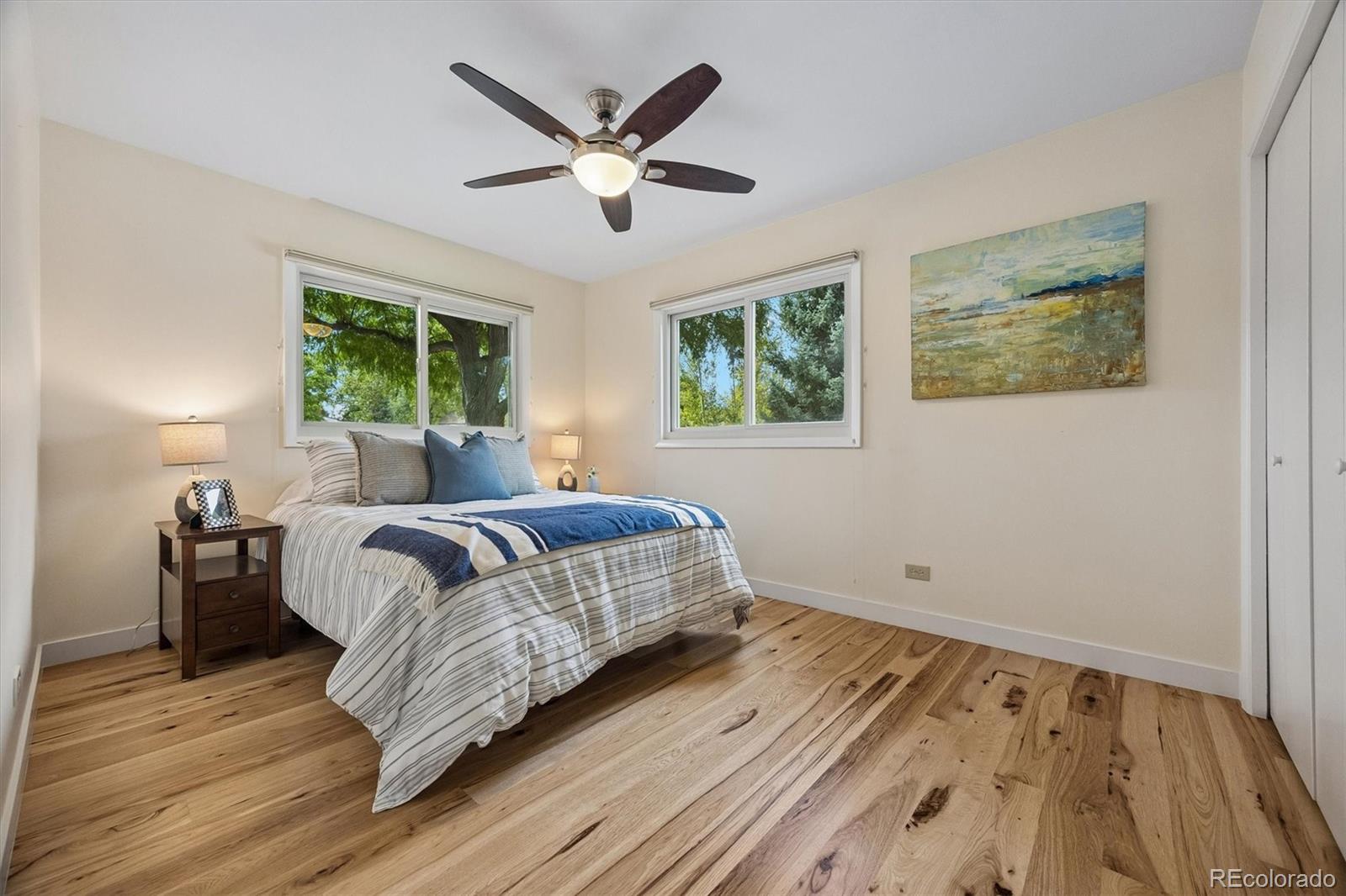 Bedroom with blue-and-white bedding, hardwood floors, a ceiling fan, and windows overlooking leafy trees.