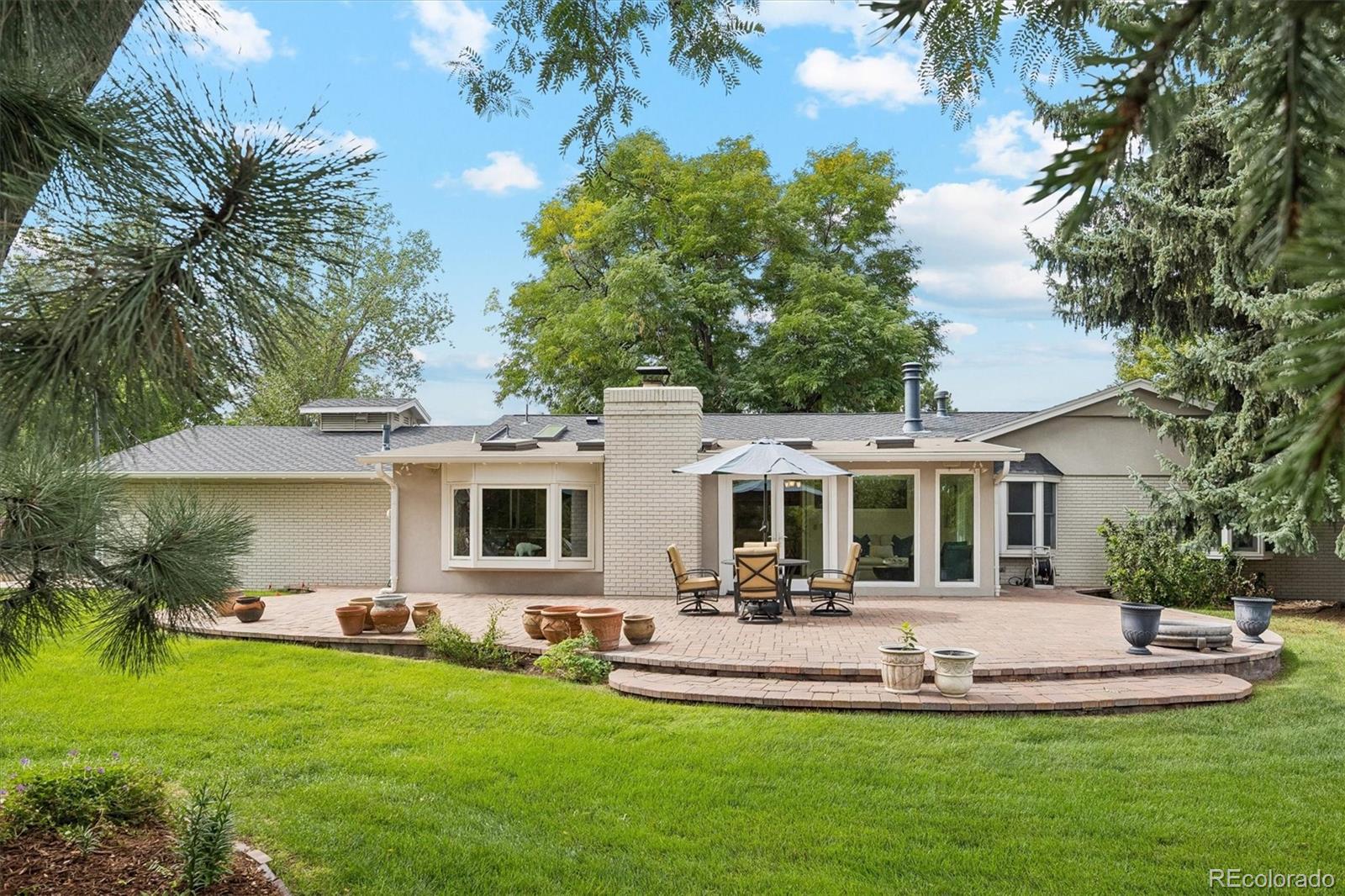 Single-story home with a broad brick patio, umbrella-shaded dining set, and mature trees.