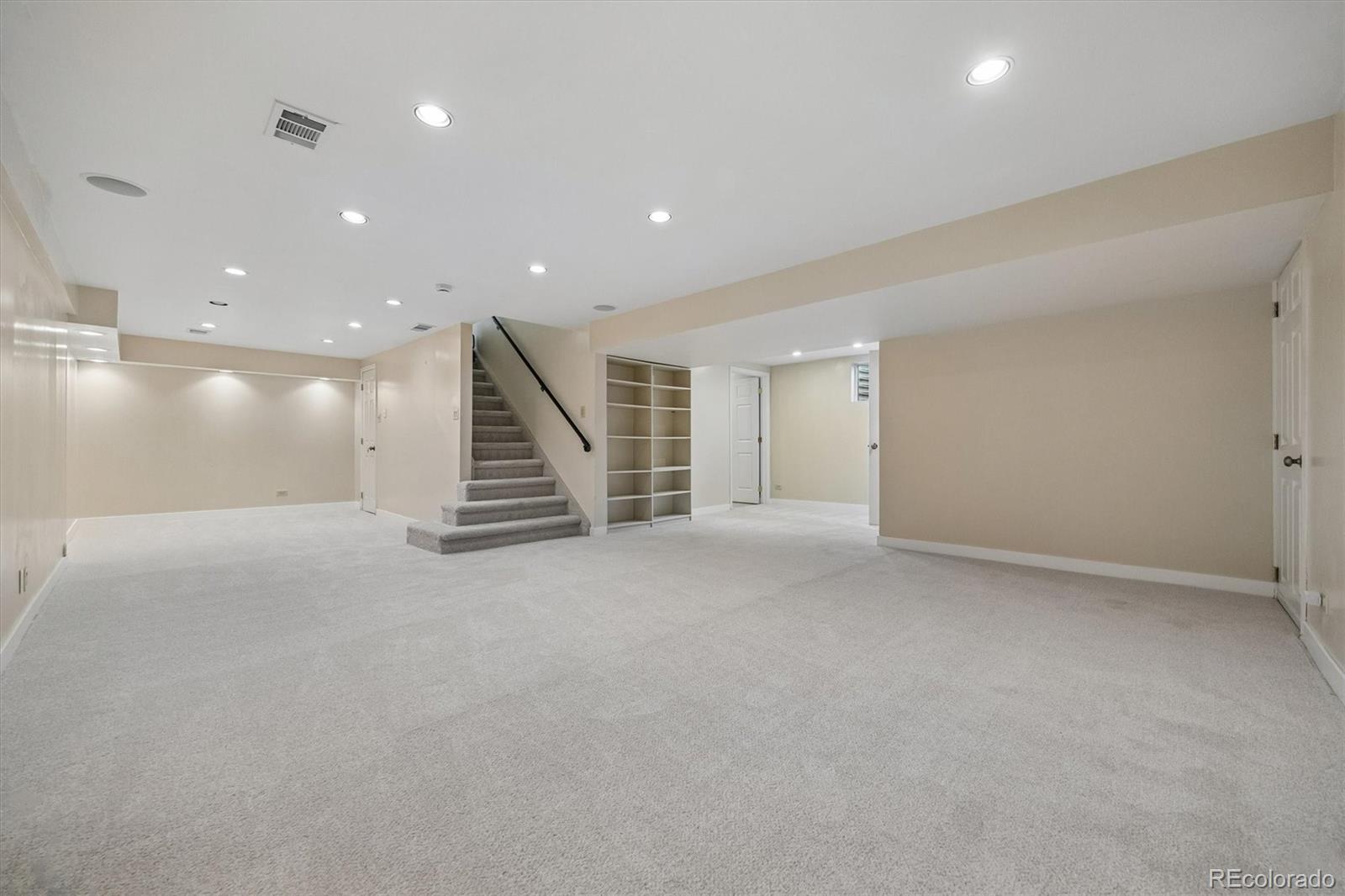 Empty carpeted basement with a staircase, dark handrail, and built-in shelving.