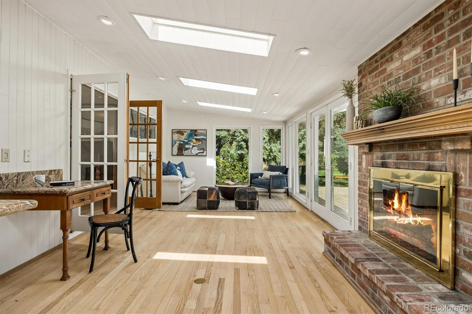 Bright sunroom with a brick fireplace, skylights, and tall windows overlooking greenery.