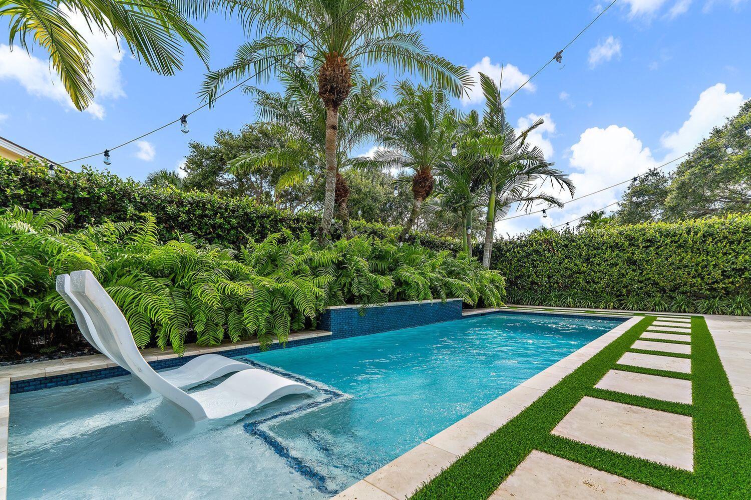 Backyard pool with built-in white loungers, tropical landscaping, and turf-lined stepping stones.