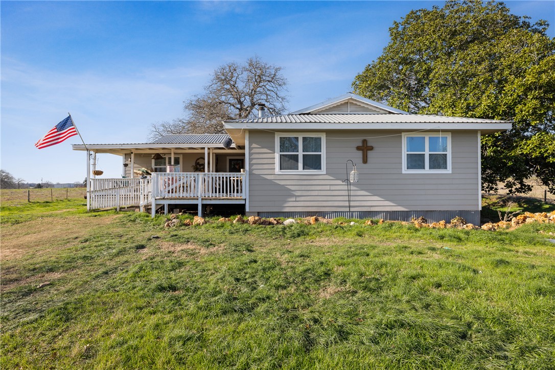 Gray ranch-style home with a covered porch, American flag, and grassy rural yard.