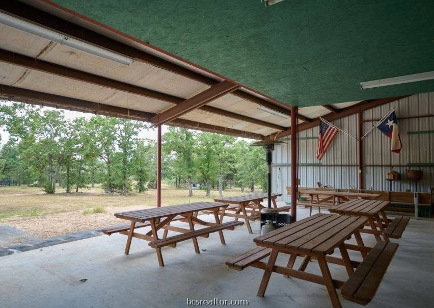 Covered pavilion with wooden picnic tables, American and Texas flags, and a tree-lined yard.