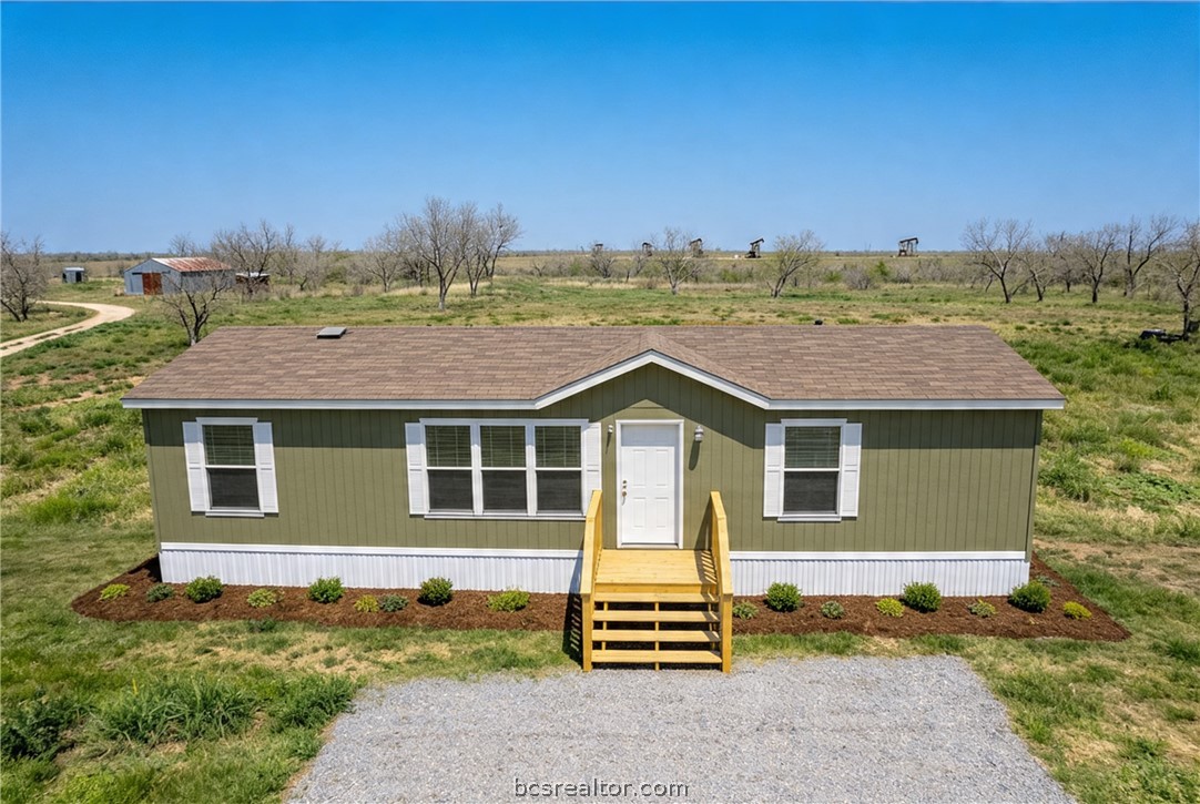 Green single-story home with wooden front steps, gravel driveway, and open rural land.