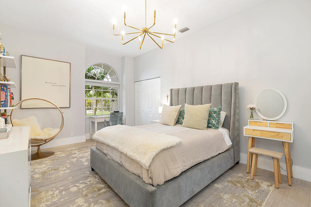 Staged bedroom with a gray upholstered bed, arched window, gold chandelier, and compact vanity.