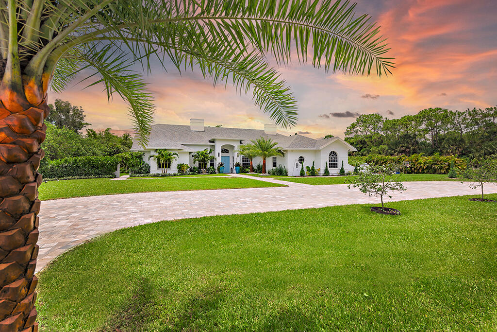White single-story home with arched windows, a broad paved driveway, and a palm tree at sunset.