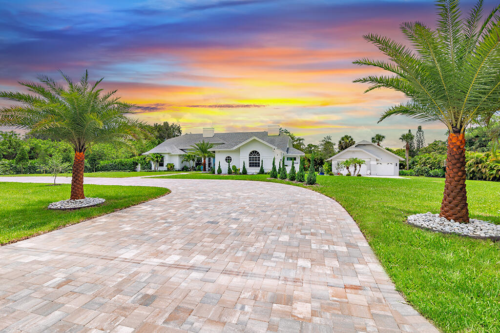 White single-story home with detached garage, curved paver driveway, and palm trees at sunset.