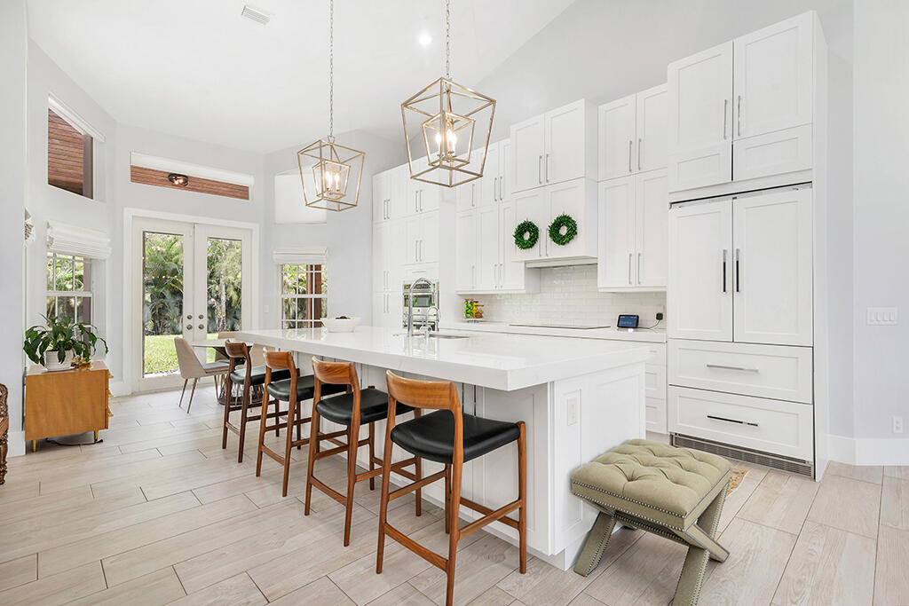 Bright white kitchen with a large island, wooden stools, and glass doors to a leafy yard.