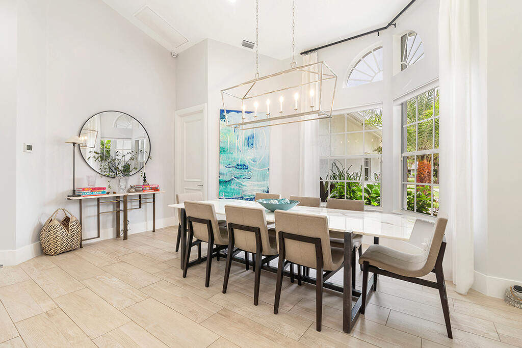 Dining room with a long white table, geometric chandelier, arched windows, and views of tropical greenery.