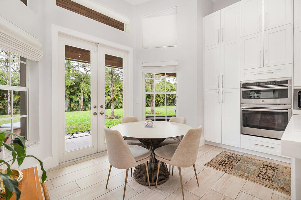 Kitchen dining nook with a round table, white cabinets, double ovens, and doors opening to a palm-lined lawn.