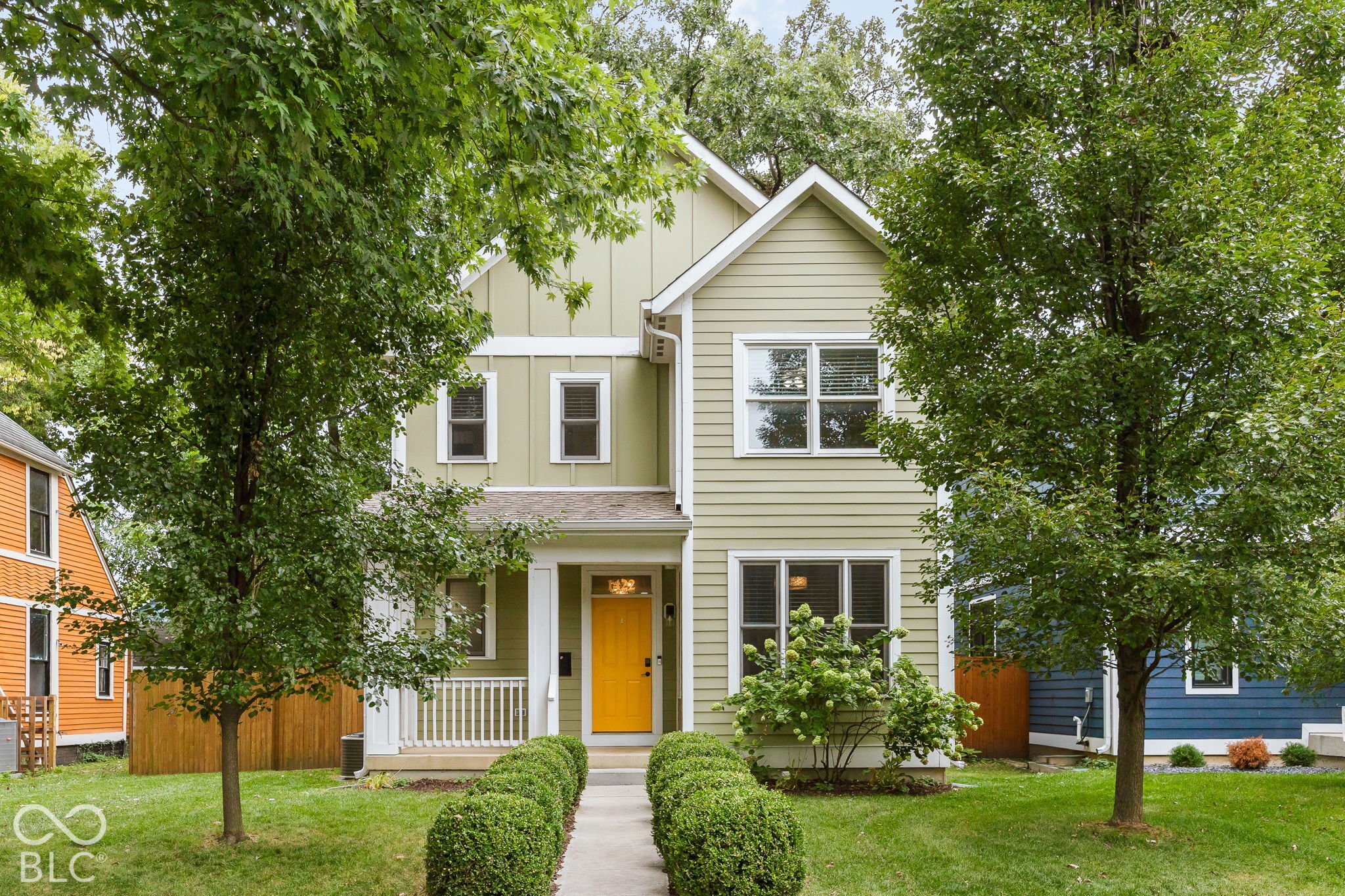Pale green two-story home with a bright yellow front door, covered porch, and leafy front yard.