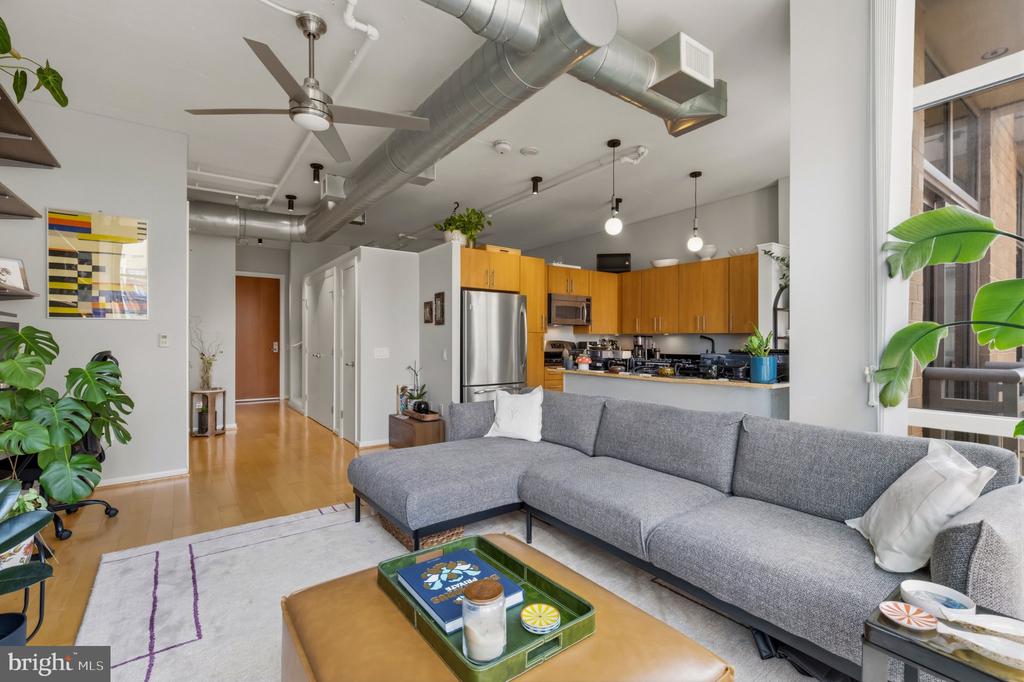 Open-plan condo living room with a gray sectional, wood kitchen cabinets, and exposed ductwork.