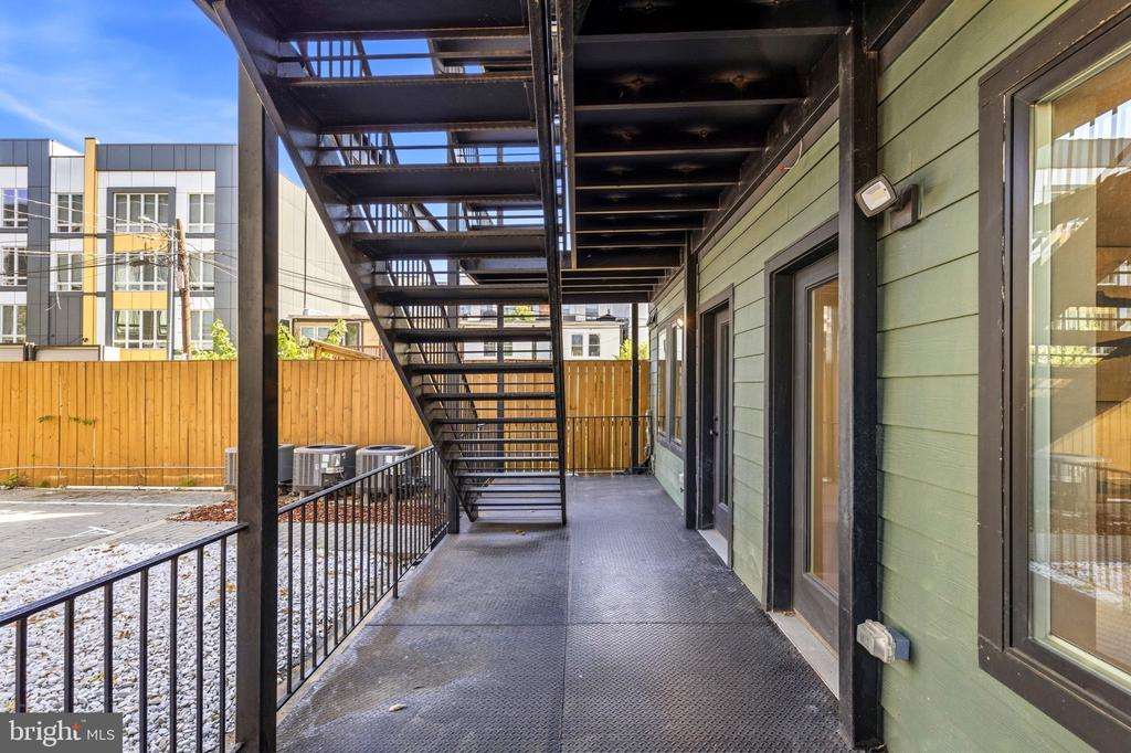 Exterior walkway beside a green-sided building, with black railings and an overhead metal staircase.