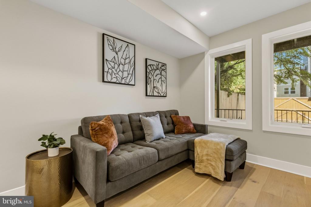 Staged living room with a gray sectional, botanical wall art, and large windows.