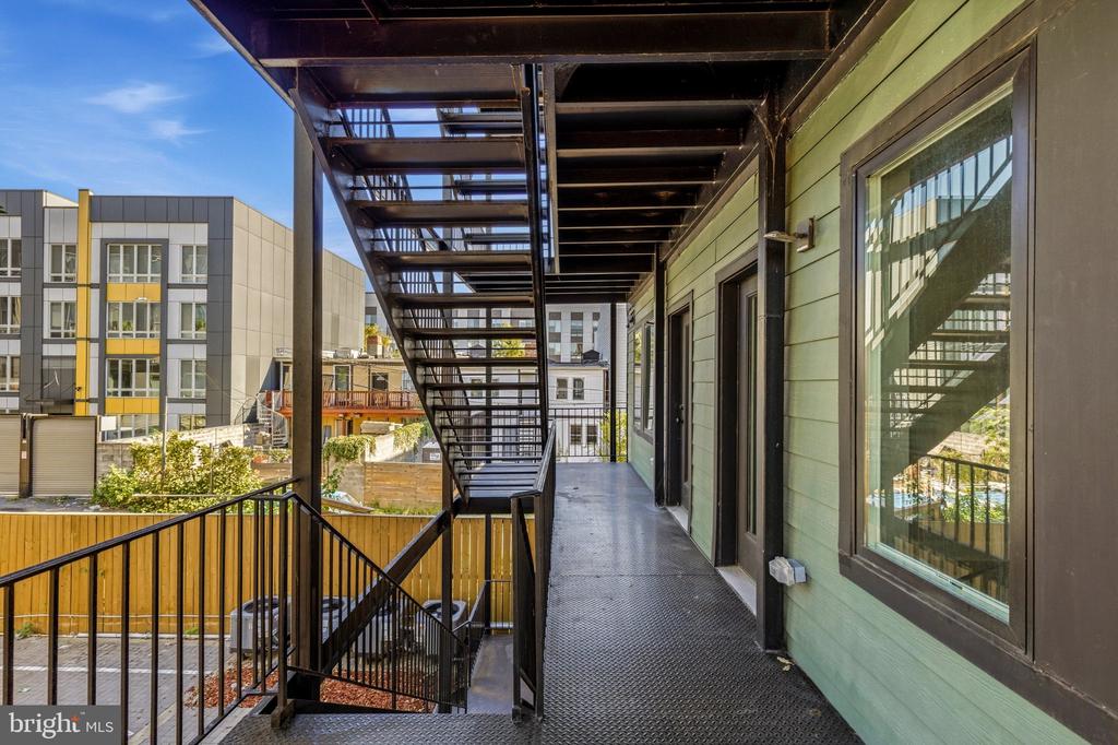 Elevated exterior walkway beside green siding, with black metal stairs and neighboring apartment buildings.