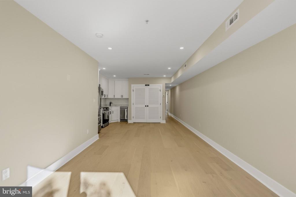 Open living area with light wood floors leading to a compact kitchen with white cabinets and stainless steel appliances.
