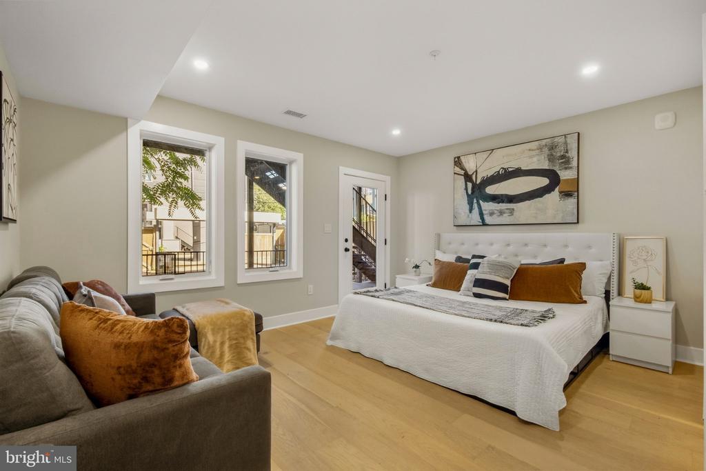 Staged bedroom with a white bed, rust-colored pillows, and a gray sofa beside two tall windows.