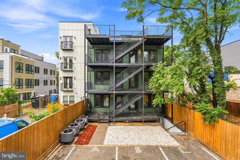 Modern multi-unit building with metal balconies and exterior stairs behind a fenced parking area.