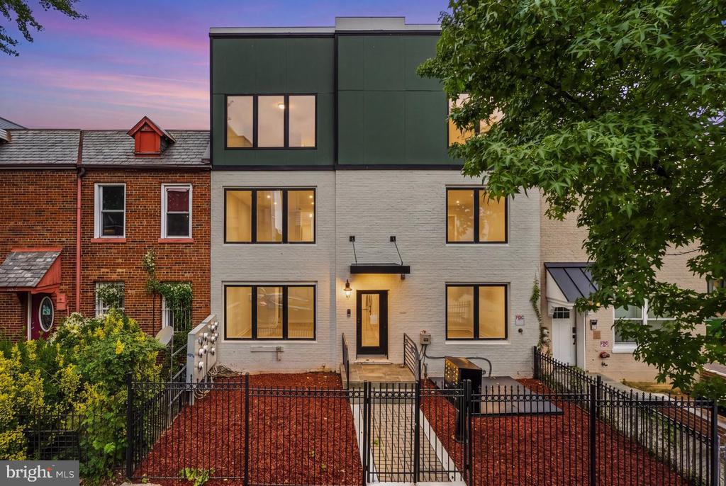 Modern townhouse with a fenced front yard, photographed at dusk between brick rowhouses.