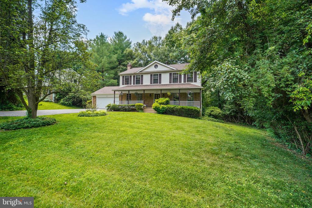 Two-story home with a covered porch and attached garage, surrounded by trees and a broad green lawn.