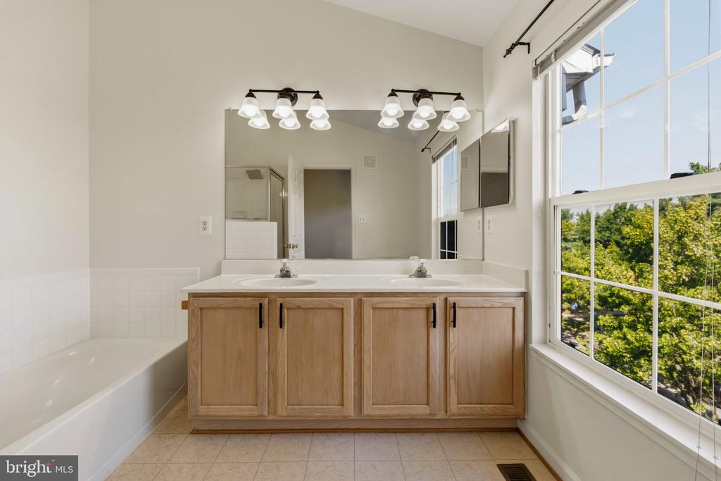 Bathroom with a double-sink wood vanity, large mirror, bathtub, and window overlooking trees.