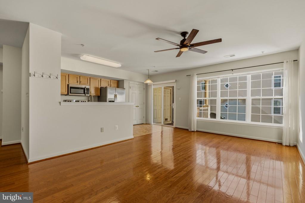 Vacant living room with polished hardwood floors, a ceiling fan, and a large window beside the open kitchen.