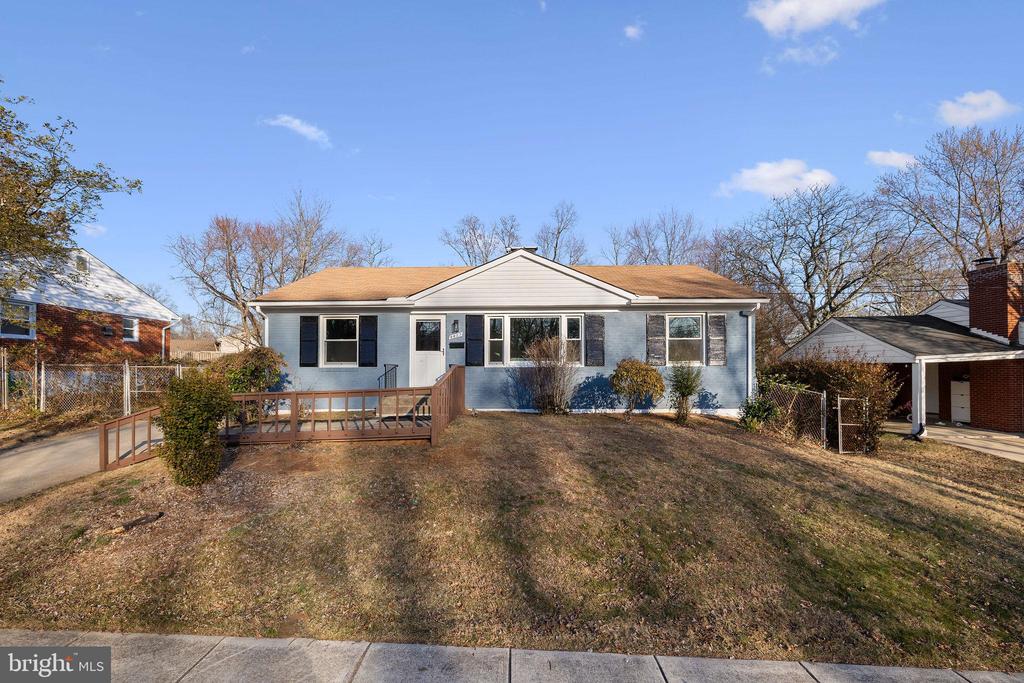Single-story blue house with a tan roof and wooden ramp across a sloped front yard.