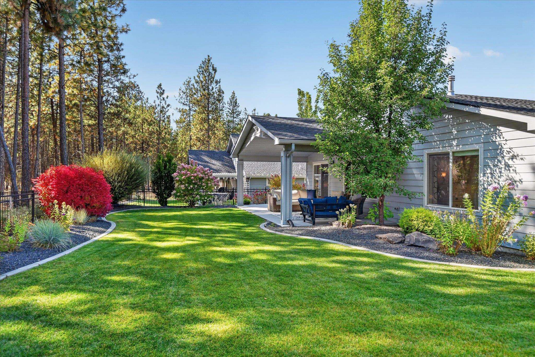 Landscaped backyard with a broad lawn, colorful garden beds, and a covered patio with outdoor seating.