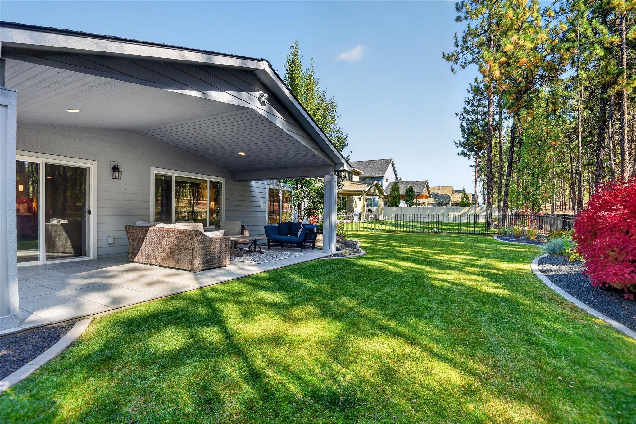 Backyard with a covered, furnished patio, broad lawn, black fencing, and mature trees.