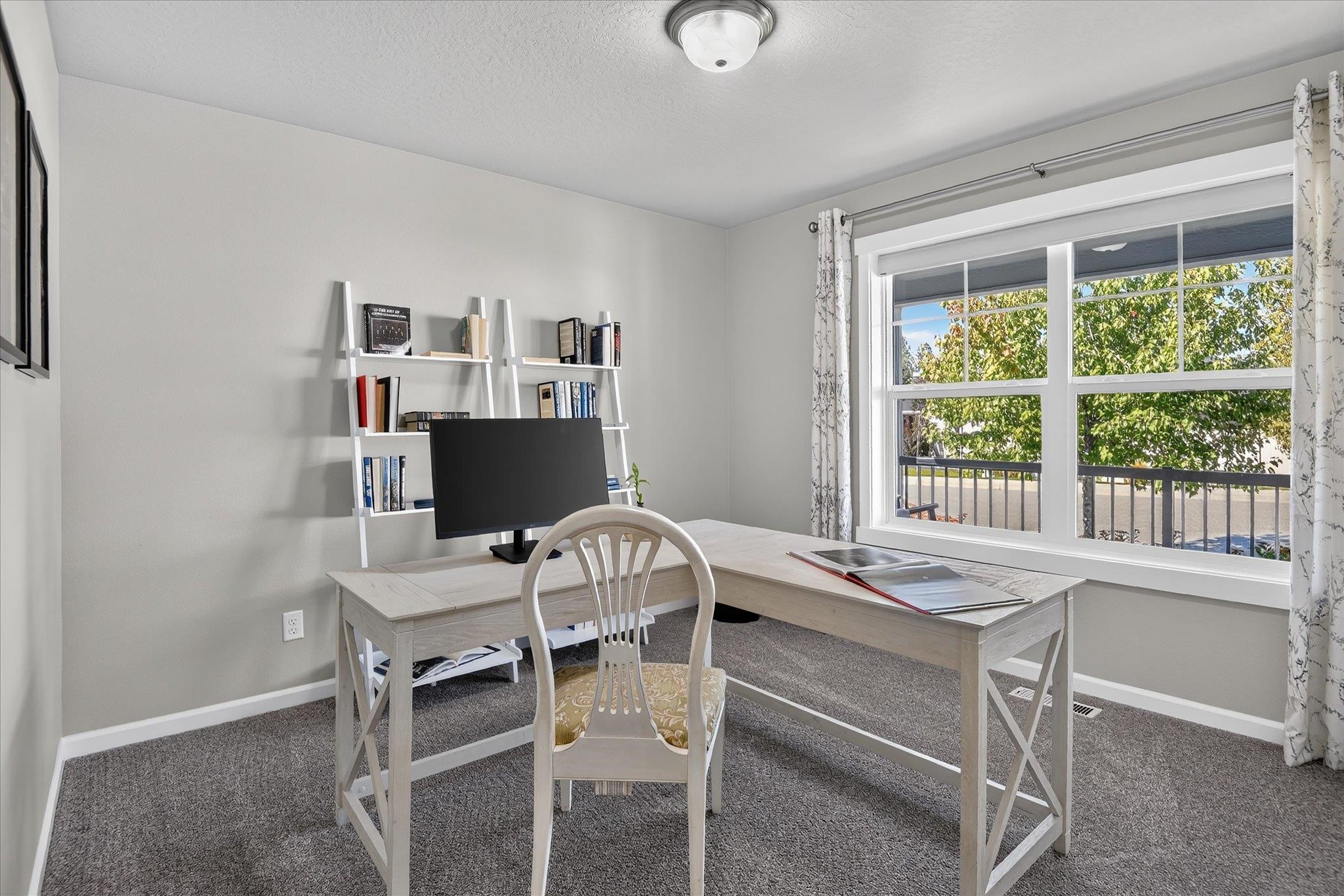 Furnished home office with an L-shaped desk and large window overlooking leafy trees.
