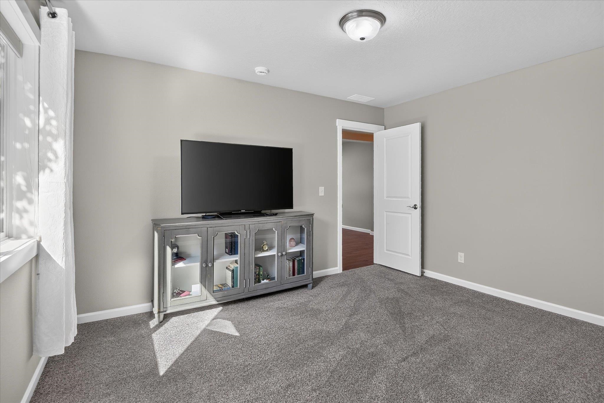 Carpeted room with a television on a glass-front cabinet beside a sunlit window.