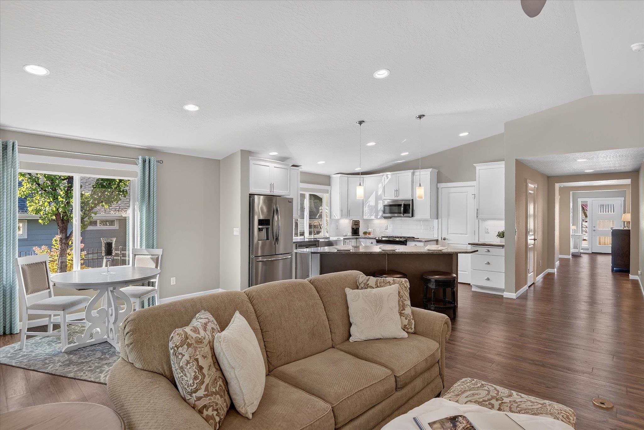 Open-plan living room with tan sofa, white kitchen, and dining nook by a large window.